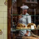 Chef behind a bakery window showcasing gourmet breads on glass shelves.